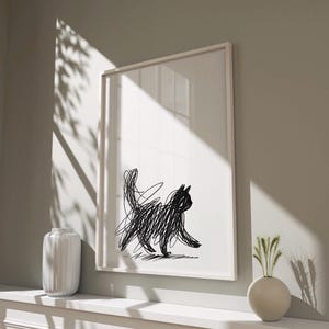 Könnte beinhalten: Ein gerahmter Kunstdruck mit einer Illustration einer schwarzen Katze. Die Katze ist in einem gekritzelten Stil auf weißem Hintergrund dargestellt. Das Kunstwerk wird in einem weißen Rahmen präsentiert, was seine moderne Ästhetik unterstreicht. Der Druck wird auf einem weißen Regal ausgestellt.