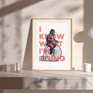 May include: A framed art print featuring a gorilla in a red suit and bowler hat riding a penny-farthing bicycle. The print has the text "I KNOW WHAT I'M DOING" in red. The artwork is displayed on a white desk with a lamp and a mug.