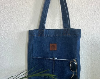 May include: A blue denim tote bag with a brown leather patch and a front pocket. Sunglasses are tucked into the pocket.