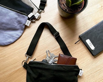 May include: Three canvas bags with adjustable straps, one black, one grey, and one beige. The black bag is open and has a brown wallet and keys inside. The bags have a white label with the word "Mia" on them.