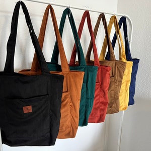 Könnte beinhalten: Ein Set aus sieben Einkaufstaschen in verschiedenen Farben, darunter Schwarz, Braun, Grün, Rot, Gelb und Blau. Die Taschen sind aus Cord und haben eine kleine Tasche auf der Vorderseite.