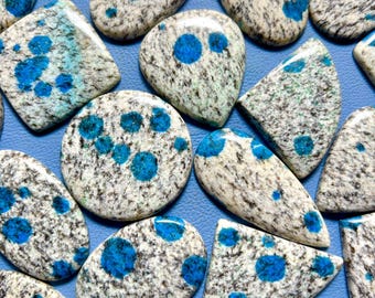 May include: A collection of K2 stone cabochons in various shapes, including ovals, teardrops, and triangles. The stones feature a speckled white and gray base with vibrant blue azurite inclusions. The cabochons are arranged on a blue surface.