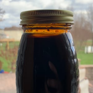 May include: A glass jar with a gold lid filled with a dark liquid. The jar has a textured surface and is sitting on a white windowsill.