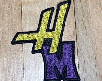Puede incluir: Un parche bordado amarillo y morado con las letras "#M" en una fuente estilizada.