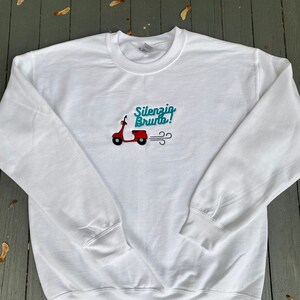 K&ouml;nnte beinhalten: Wei&szlig;er Pullover mit einem roten Roller und dem gestickten Text "Silenzio Bruto!" auf der Vorderseite.