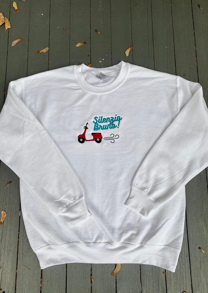 K&ouml;nnte beinhalten: Wei&szlig;er Pullover mit einem roten Roller und dem gestickten Text "Silenzio Bruto!" auf der Vorderseite.