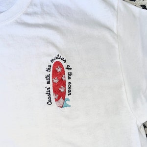 May include: White t-shirt with red and blue embroidered surfboard and the text "Coastin' with the motion of the ocean".