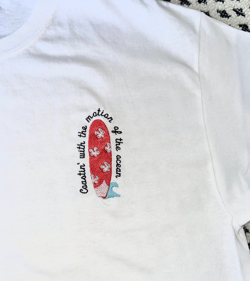 May include: White t-shirt with red and blue embroidered surfboard and the text "Coastin' with the motion of the ocean".