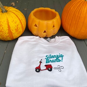 K&ouml;nnte beinhalten: Wei&szlig;es Sweatshirt mit einem t&uuml;rkisfarbenen gestickten Design eines roten Rollers mit dem Text "Silenzio Bruto!" auf einem Holzgrund mit zwei K&uuml;rbissen.