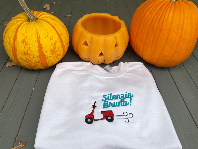 K&ouml;nnte beinhalten: Wei&szlig;es Sweatshirt mit einem t&uuml;rkisfarbenen gestickten Design eines roten Rollers mit dem Text "Silenzio Bruto!" auf einem Holzgrund mit zwei K&uuml;rbissen.