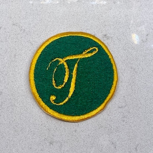 May include: Circular embroidered patch with a green centre and a gold border. A gold, cursive letter "T" is embroidered in the middle. Suitable for application on clothing or accessories.