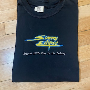 May include: Black t-shirt with a yellow and blue embroidered logo that says "Sonny Eclipse" and the text "Biggest Little Star in the Galaxy". The shirt has a tag that says "Comfort Colors".