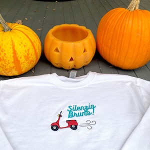 K&ouml;nnte beinhalten: Wei&szlig;es Sweatshirt mit einem roten Roller-Motiv und dem Text "Silenzio Brunto!" in t&uuml;rkisfarbener Schrift. Das Sweatshirt liegt auf einer Holzoberfl&auml;che mit zwei orangefarbenen K&uuml;rbissen.