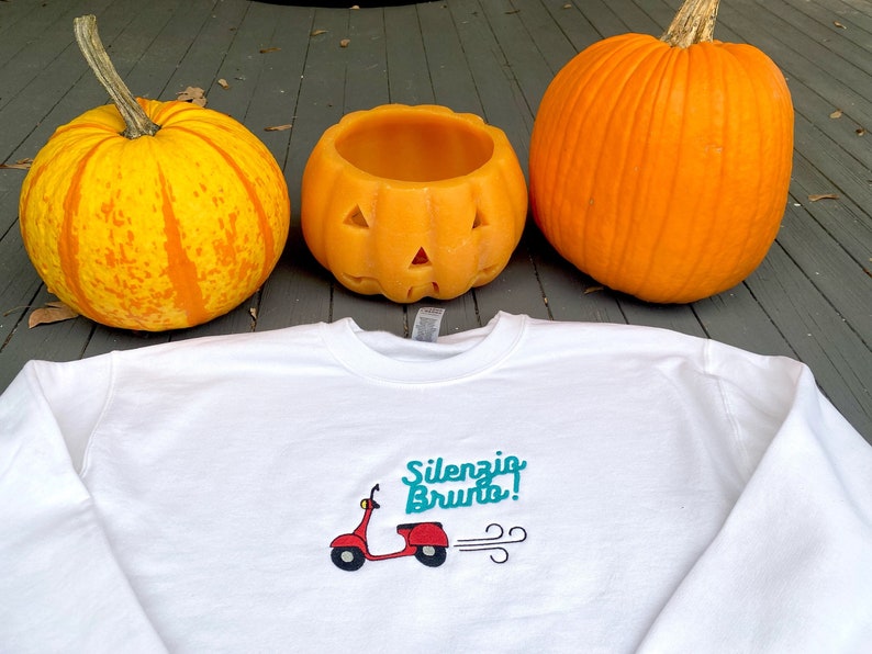 K&ouml;nnte beinhalten: Wei&szlig;es Sweatshirt mit einem roten Roller-Motiv und dem Text "Silenzio Brunto!" in t&uuml;rkisfarbener Schrift. Das Sweatshirt liegt auf einer Holzoberfl&auml;che mit zwei orangefarbenen K&uuml;rbissen.