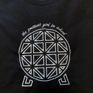Pu&ograve; includere: Maglietta nera con ricamo bianco di un disegno geometrico e il testo "the prettiest girl in school".