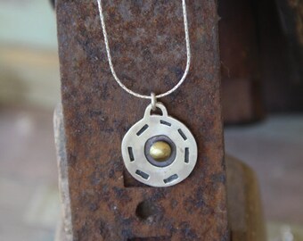 Puede incluir: Collar con colgante de plata con un diseño circular. El colgante tiene un anillo exterior plateado con detalles recortados y un centro dorado. El collar se muestra sobre una superficie marrón rústica.