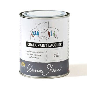 May include: A white can of Annie Sloan Chalk Paint Lacquer in clear gloss. The can has a hand-painted design of a person with colorful fingertips. The text on the can reads "Chalk Paint Lacquer", "a hard-wearing varnish for both interiors and exteriors", "Clear Gloss", and "UV Protection".