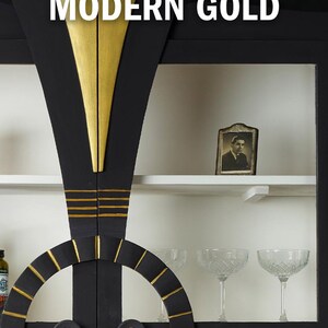 May include: A black cabinet with gold accents. The cabinet has two doors with a circular design and a gold trim. The doors are closed. The cabinet has shelves with a white background. There is a small framed photo on the top shelf. The cabinet is labeled "MODERN GOLD".
