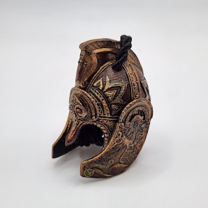 Puede incluir: Un casco detallado de color bronce con intrincadas tallas y una cuerda negra adjunta. El casco presenta un diseño de cabeza de caballo en la parte superior y los paneles laterales. El diseño general es ornamentado e histórico.