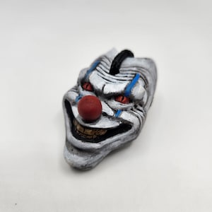 Puede incluir: Figurita de cabeza de payaso pintada a mano. El payaso tiene una cara blanca con detalles negros y azules, ojos rojos y una nariz roja. La boca está abierta, revelando dientes amarillos. Una cuerda negra está unida a la parte superior de la cabeza.