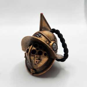 Puede incluir: Un casco de gladiador de color dorado con una correa trenzada negra. El casco tiene una cresta elevada y una máscara facial con agujeros.
