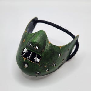 Puede incluir: Una máscara de metal verde con un acabado desgastado, con una abertura para la boca con barras metálicas y numerosos agujeros pequeños. Una cuerda negra está unida para asegurar la máscara.