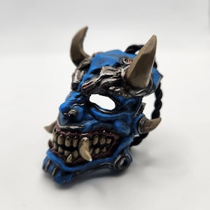 May include: A detailed blue and brown Oni mask with sharp teeth, horns, and intricate detailing. The mask features a menacing expression and is designed with a textured finish, showcasing a blend of blue and brown hues.