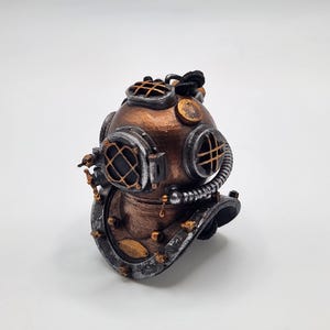 Könnte beinhalten: Ein kleiner, detaillierter Taucherhelm aus Kupfer und Silber mit einem schwarzen Band. Der Helm hat eine runde Form mit einem klaren Glasfenster und einem dekorativen Design.