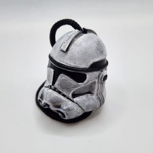 May include: A silver and black helmet ornament, resembling a Star Wars Clone Trooper helmet. The helmet has a textured, weathered appearance with a black trim and a black loop for hanging. The ornament is set against a white background.