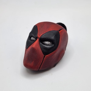 May include: A red and black Deadpool mask with a textured surface. The mask features black eye coverings with mesh detailing and a black head covering.