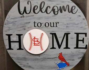 May include: A round wooden sign with a gray distressed paint finish. The sign says "Welcome to our HOME" with a baseball with the St. Louis Cardinals logo in the center. A red cardinal bird is perched on a blue musical note.