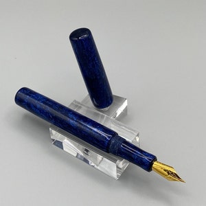 Puede incluir: Una pluma estilogr&aacute;fica azul y dorada con un cuerpo marmoleado azul y una plumilla dorada. La pluma se muestra en un soporte de acr&iacute;lico transparente.