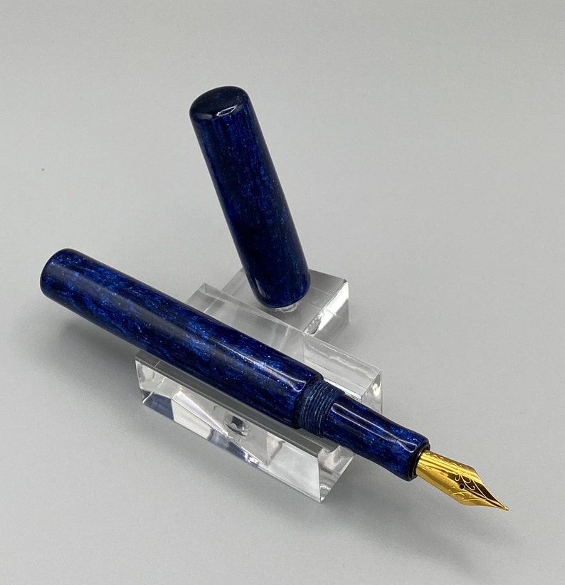 Puede incluir: Una pluma estilogr&aacute;fica azul y dorada con un cuerpo marmoleado azul y una plumilla dorada. La pluma se muestra en un soporte de acr&iacute;lico transparente.