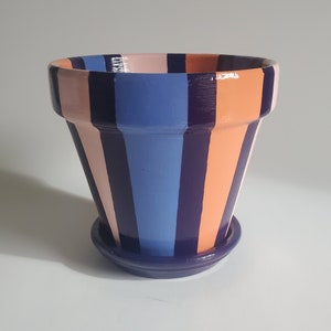 Op de afbeelding: Een terracotta plantenpot met een gestreept ontwerp in tinten blauw, roze en paars. De pot heeft een bijpassende schotel.