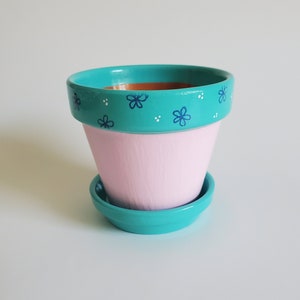 May include: A pink terracotta planter with a turquoise rim and painted blue flowers. The planter has a matching turquoise saucer.