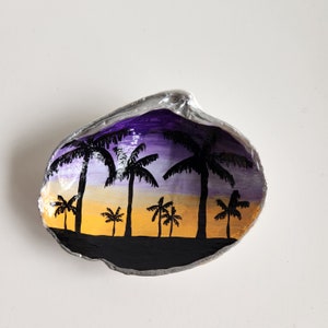 May include: A seashell painted with a sunset scene featuring palm trees in silhouette against a purple and yellow sky.