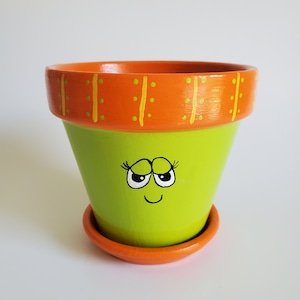 May include: A green terracotta planter with a painted orange rim and a cute cartoon face. The face has black eyelashes, white eyes, and a black smile.