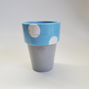 May include: A small, hand-painted flower pot. The pot is gray with a light blue rim. White polka dots are painted on the blue rim. The pot is empty and ready for planting.