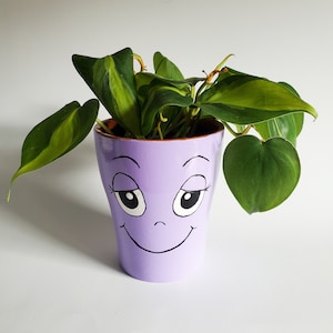 May include: A purple ceramic planter with a painted smiley face and large eyes. The planter is filled with a green leafy plant.