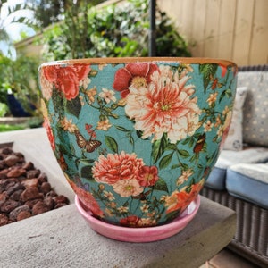 Könnte beinhalten: Ein türkisfarbener Blumentopf mit einem floralen Muster mit rosa, weißen und roten Blumen und Schmetterlingen. Der Topf hat einen rosa Untersetzer.