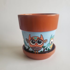 May include: A terracotta flower pot with a blue background and a hand-painted design of three owls. The owls are orange, blue, and green with black eyes and white pupils. The pot has a brown rim and a brown saucer.