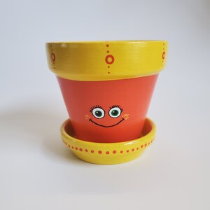 May include: A yellow and orange terracotta planter with a painted smiley face and a matching saucer. The planter has red dots and circles around the rim.