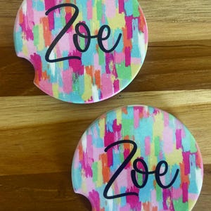 May include: Two round car coasters with a colorful abstract design in pink, blue, yellow, and orange. The word "Zoe" is written in black cursive on each coaster. The coasters have a curved cutout for easy removal.