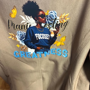 May include: A tan jacket featuring a graphic print of a person with dark hair wearing sunglasses and a blue sweatshirt that says FOCUSED. The print includes yellow butterflies, blue roses, and the text Manifesting GREATNESS in black and blue.
