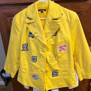 May include: A bright yellow blazer featuring three-quarter sleeves, a notched collar with black bead-adorned safety pins, and white pearl accents. The jacket is decorated with various embroidered patches displaying text including Faith, Yes You Can, and Dream It.