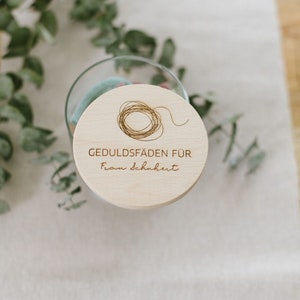 Könnte beinhalten: Ein Holzdeckel mit einem Kreis aus Schnur und dem eingravierten Text "Geduld Fäden für Frau Schubert".