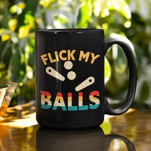 May include: Black ceramic mug with a white and yellow graphic that says "Flick My Balls" with a retro pinball design.