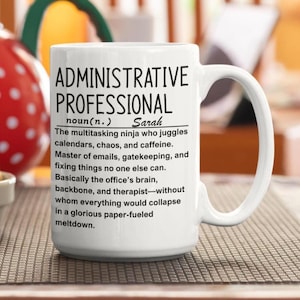 Op de afbeelding: Een witte keramische mok met zwarte tekst die een administratieve professional definieert als een multitasking ninja die jongleert met agenda's, chaos en cafeïne, en fungeert als het brein, de ruggengraat en de therapeut van het kantoor.