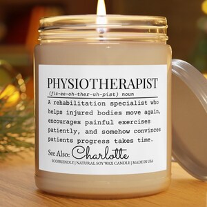 Puede incluir: Una vela beige encendida en un tarro de cristal con una etiqueta blanca. La etiqueta dice "PHYSIOTHERAPIST" con una definición y el nombre "Charlotte". La vela está hecha de cera de soja natural.