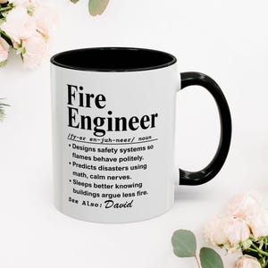 Op de afbeelding: Een witte keramische mok met een zwarte binnenkant en handvat. De mok heeft de tekst "Fire Engineer" met een definitie, inclusief de uitspraak en een lijst met taken. De tekst "See Also: David" is ook opgenomen.